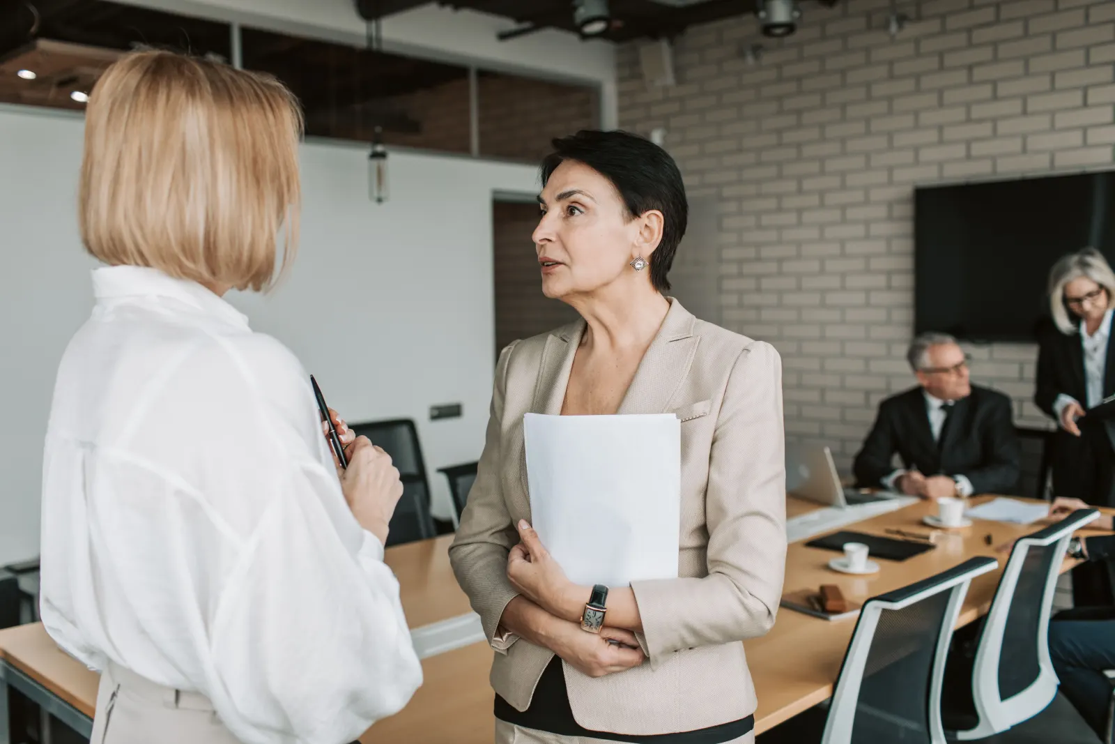 Two managers discussing a project in a meeting room while colleagues work at the table behind them